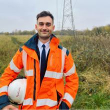 Male engineer wearing a high-viz safety jacket in a field setting, in front of a large electricity pylon